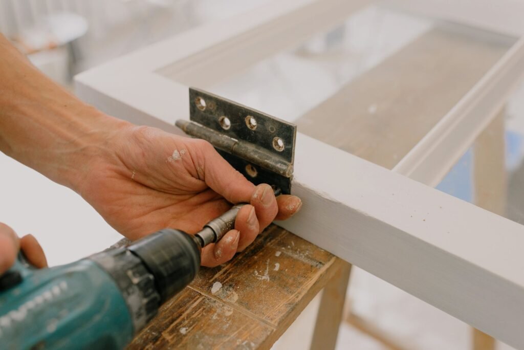 Primer plano de las manos de un manitas utilizando un taladro para fijar una bisagra en el marco de una ventana.