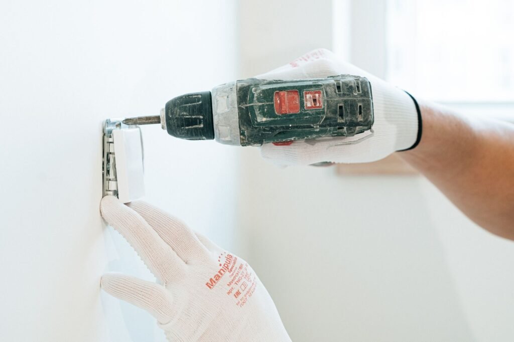 Manos con guantes usando un taladro para instalar un enchufe en la pared, enfocándose en la precisión y la seguridad.