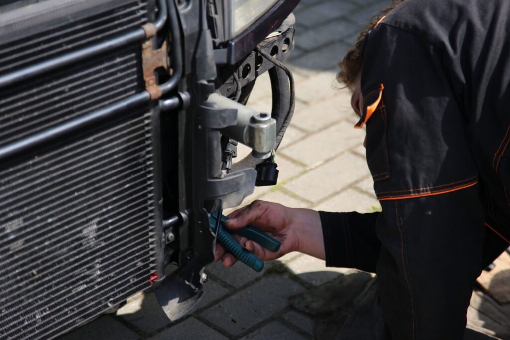 Mecánico trabajando en la reparación de coches al aire libre utilizando herramientas, enfocándose en el área del radiador del vehículo.