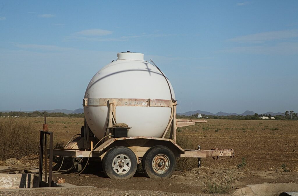 Un tanque de agua redondo sobre un remolque, ubicado en un campo rural bajo un cielo azul despejado.