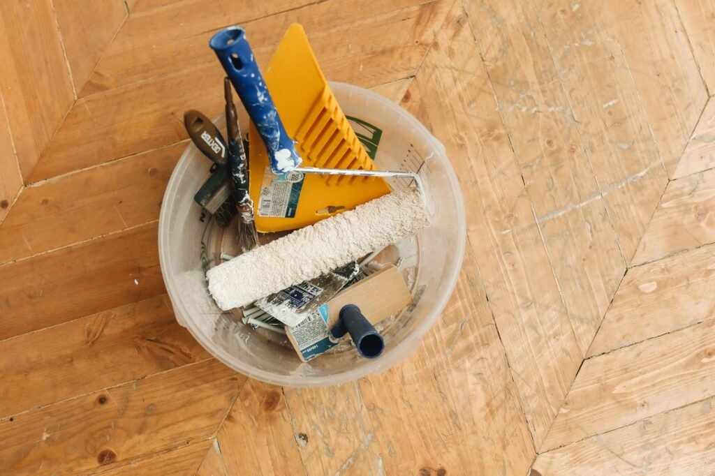 Una colección de herramientas de renovación del hogar en un balde sobre un suelo de madera, listas para un proyecto de pintura.